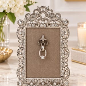 May include: A rectangular picture frame with an ornate, silver-toned metal border encrusted with clear crystals. The frame features a central taupe fabric backing and a decorative silver-toned metal ornament with a hanging ring at the center.