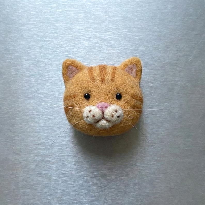 Pu&ograve; includere: Un volto di gatto tabby arancione fatto a mano in feltro. Il gatto ha occhi neri, naso rosa e baffi. Il pelo del gatto presenta strisce arancioni pi&ugrave; scure. Un oggetto decorativo.