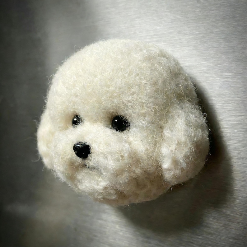 May include: A white, fluffy, felted dog head with black eyes and nose. The dog's fur is textured and detailed, giving it a realistic appearance. The dog's head is round and has a friendly expression.