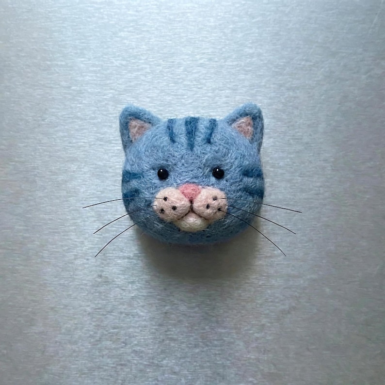 Pu&ograve; includere: Testa di gatto in feltro blu con strisce blu scuro, interno orecchie rosa e naso rosa. Il gatto ha occhi neri e lunghi baffi neri. &Egrave; un oggetto fatto a mano.