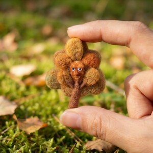 K&ouml;nnte beinhalten: Ein handgefertigter Filzbaum mit einem skurrilen Gesicht. Der Baum hat einen braunen Stamm und &Auml;ste mit orangefarbenen und gr&uuml;nen Bl&auml;ttern. Der Baum wird zwischen zwei Fingern vor einem verschwommenen gr&uuml;nen Hintergrund gehalten.