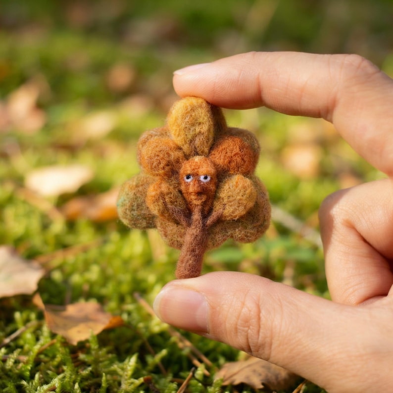 K&ouml;nnte beinhalten: Ein handgefertigter Filzbaum mit einem skurrilen Gesicht. Der Baum hat einen braunen Stamm und &Auml;ste mit orangefarbenen und gr&uuml;nen Bl&auml;ttern. Der Baum wird zwischen zwei Fingern vor einem verschwommenen gr&uuml;nen Hintergrund gehalten.
