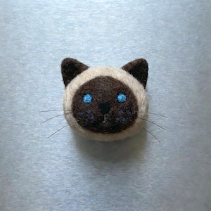 Pu&ograve; includere: Un volto di gatto siamese fatto a mano in feltro. Il gatto ha un muso e orecchie marroni, con occhi blu e baffi. Il volto del gatto &egrave; circondato da feltro color crema.