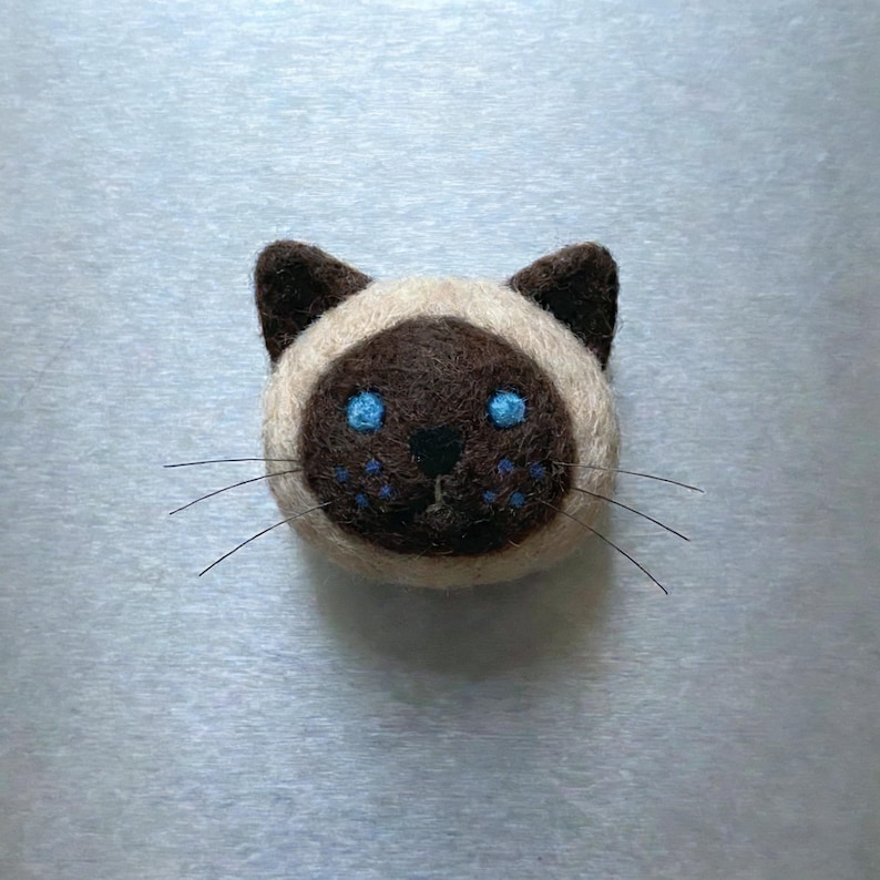 Pu&ograve; includere: Un volto di gatto siamese fatto a mano in feltro. Il gatto ha un muso e orecchie marroni, con occhi blu e baffi. Il volto del gatto &egrave; circondato da feltro color crema.