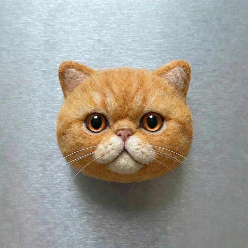 May include: A detailed, orange cat head sculpture with large, round, amber-colored eyes and a pink nose. The cat has a fluffy, textured appearance, with visible whiskers. The sculpture is likely made of felt or a similar material, and is set against a plain background.