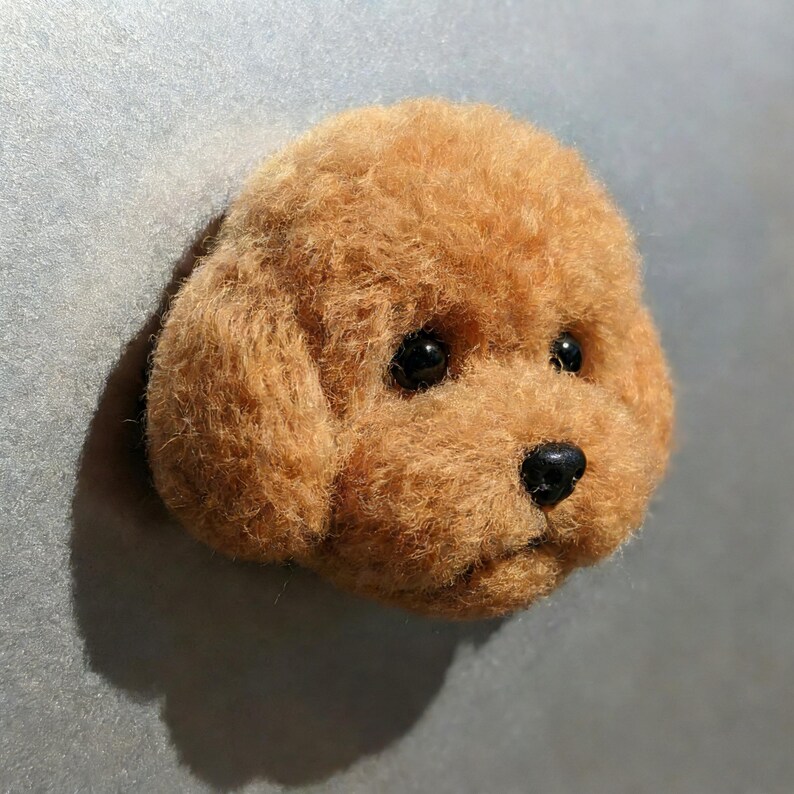 May include: A lifelike, three-dimensional sculpture of a dog's head. The dog has a fluffy, light brown coat, black eyes, and a black nose. The sculpture is set against a gray background, creating a striking contrast.