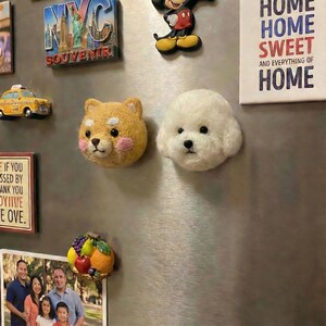 May include: A collection of decorative magnets on a refrigerator. The magnets include a yellow taxi cab, a Shiba Inu dog head, a white Bichon Frise dog head, a Mickey Mouse figure, and a NYC souvenir. A sign reads "HOME SWEET HOME".