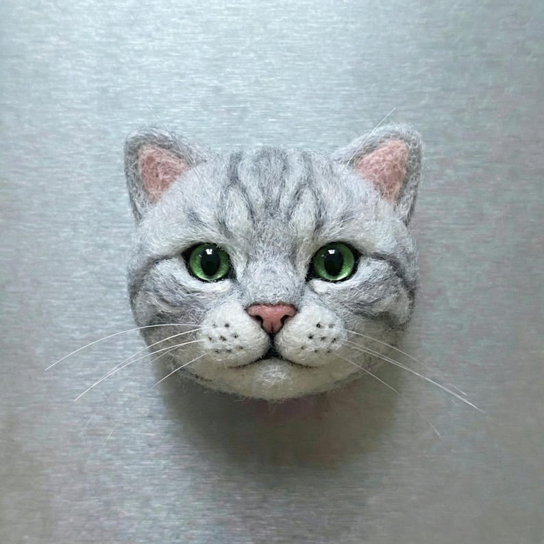 May include: A handcrafted, gray cat head with green eyes, pink nose, and detailed fur texture. The cat has long white whiskers and is centered against a light gray background. The cat's ears have pink inner ears.