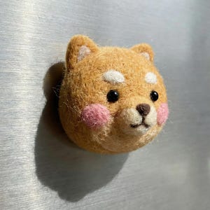 May include: A handcrafted felt Shiba Inu dog head with a light brown coat, white accents, and pink cheeks. The dog has black eyes and a brown nose. This decorative item is likely a magnet or pin.
