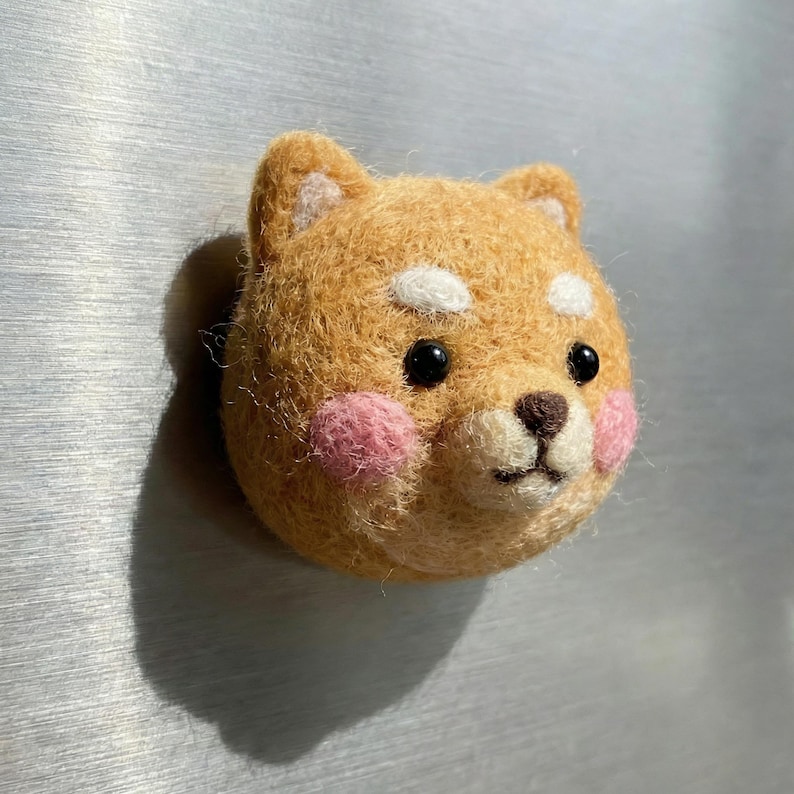 May include: A handcrafted felt Shiba Inu dog head with a light brown coat, white accents, and pink cheeks. The dog has black eyes and a brown nose. This decorative item is likely a magnet or pin.