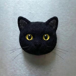 May include: A black cat head with yellow eyes and long whiskers. The cat is made of a felt-like material and is likely a decorative item or a magnet. The cat's ears are pointed, and the eyes are wide and expressive.