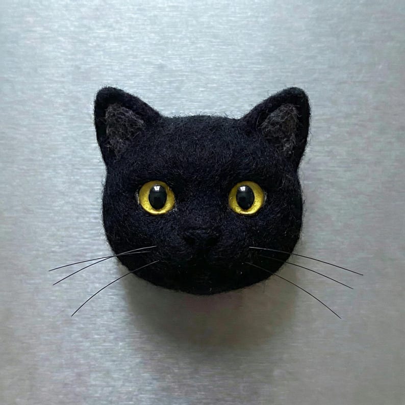May include: A black cat head with yellow eyes and long whiskers. The cat is made of a felt-like material and is likely a decorative item or a magnet. The cat's ears are pointed, and the eyes are wide and expressive.