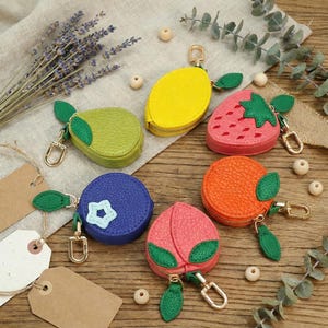 Puede incluir: Una colección de seis monederos con forma de fruta hechos de material texturizado, que incluye un limón amarillo, una pera verde, una fresa rosa, un arándano azul con una estrella, una naranja y un melocotón rosa, cada uno con un cierre de metal dorado.