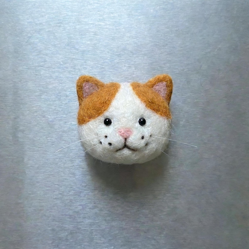 Pu&ograve; includere: Un volto di gatto in feltro fatto a mano, con pelo arancione e bianco. Il gatto ha occhi di perline nere, un naso rosa e baffi. Questo volto di gatto stravagante &egrave; un design affascinante.