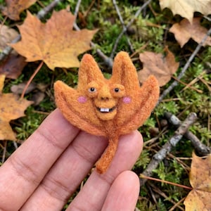 K&ouml;nnte beinhalten: Ein skurriles, orangefarbenes Filzblatt mit einem l&auml;chelnden Gesicht, rosa Wangen und kleinen wei&szlig;en Z&auml;hnen. Das Blatt hat einen Stiel und wird vor einem Hintergrund aus Herbstlaub und Gr&uuml;n gehalten. Einzigartige Herbstdekoration.