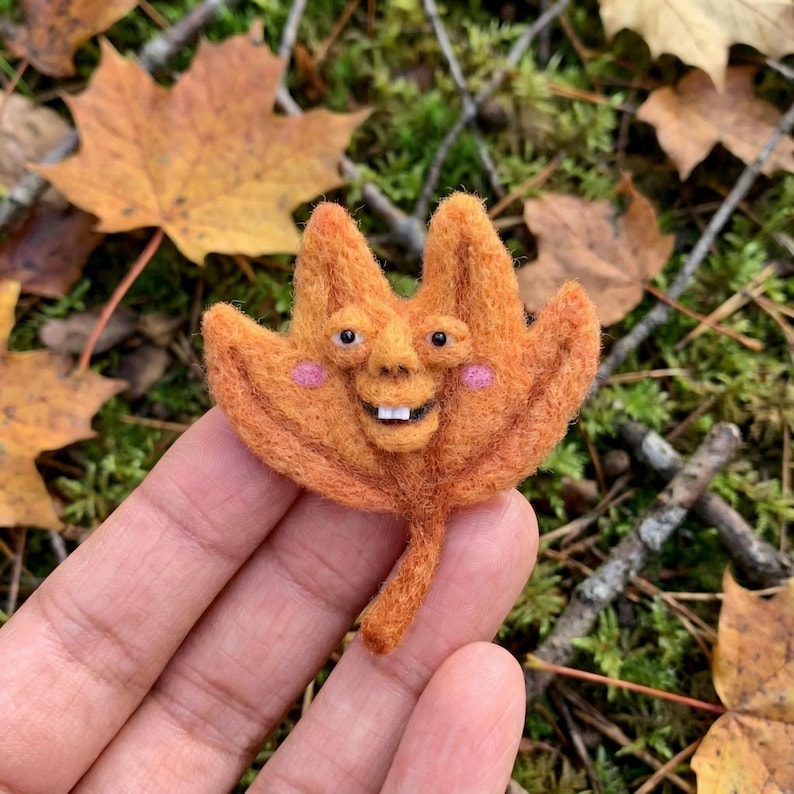 K&ouml;nnte beinhalten: Ein skurriles, orangefarbenes Filzblatt mit einem l&auml;chelnden Gesicht, rosa Wangen und kleinen wei&szlig;en Z&auml;hnen. Das Blatt hat einen Stiel und wird vor einem Hintergrund aus Herbstlaub und Gr&uuml;n gehalten. Einzigartige Herbstdekoration.