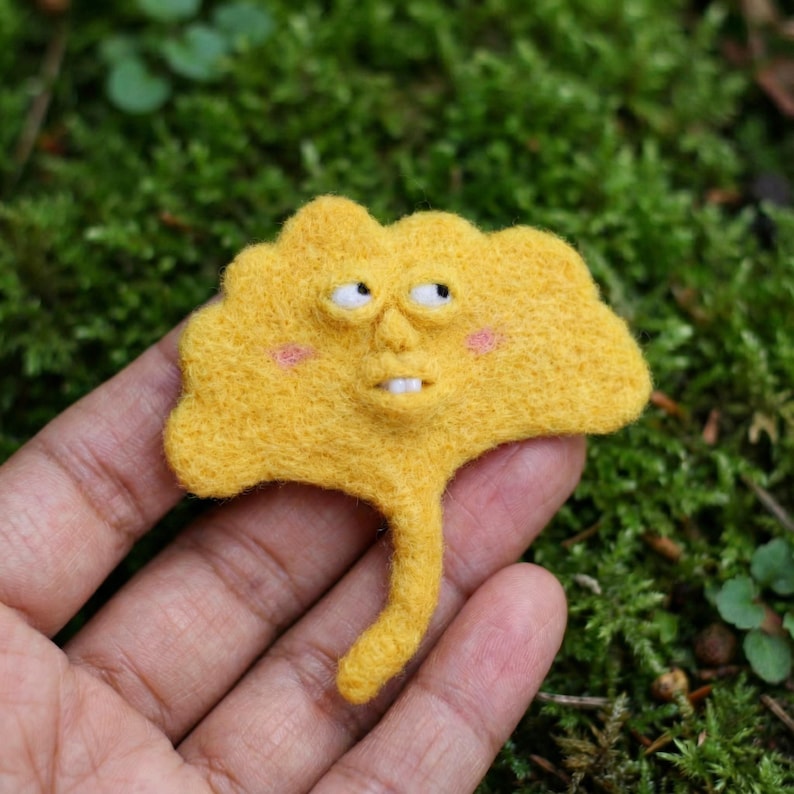 K&ouml;nnte beinhalten: Ein skurriles, handgefertigtes gelbes Filz-Ginkgoblatt mit Gesicht, komplett mit Augen, Mund und rosigen Wangen. Das Blatt hat einen Stiel und ist vor gr&uuml;nem Moos platziert. Dieser einzigartige Artikel ist ein charmantes Kunstwerk.
