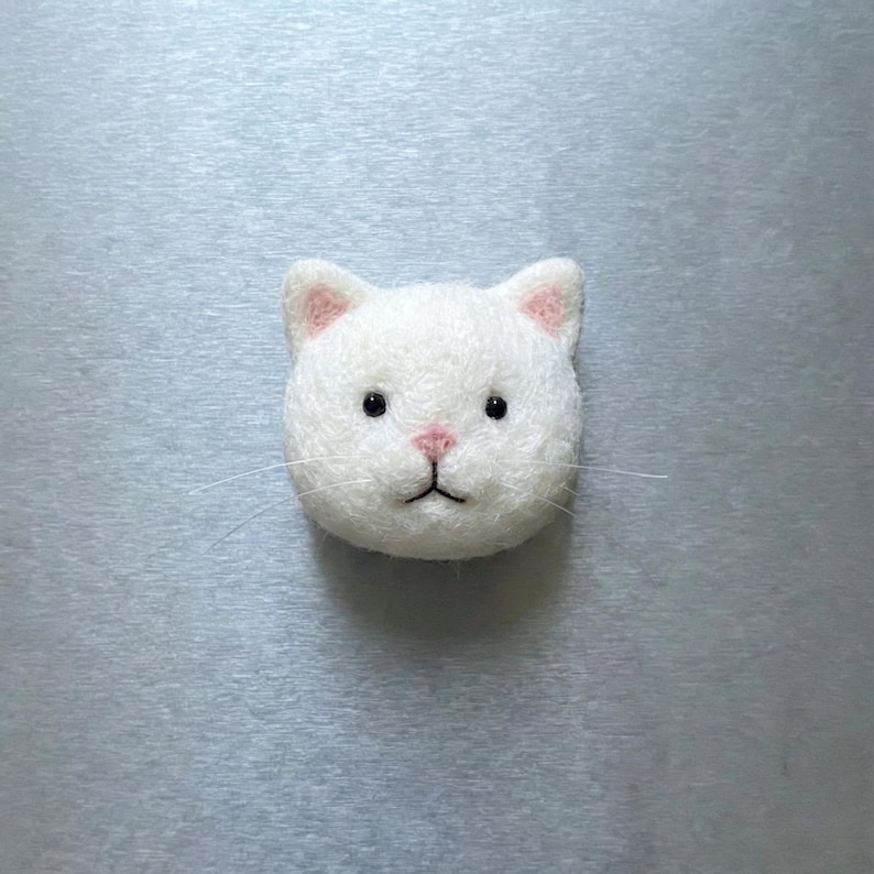 Pu&ograve; includere: Una testa di gatto in feltro bianco con l'interno delle orecchie e il naso rosa. Il gatto ha occhi neri e sottili baffi. Questo oggetto decorativo fatto a mano rappresenta un volto di gatto.