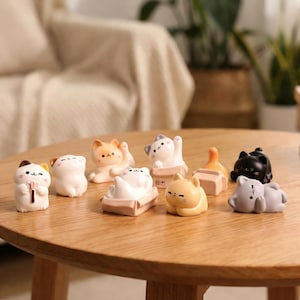 May include: A collection of small, decorative cat figurines in various poses and colors, including white, orange, black, and gray. Some cats are lying down, while others are sitting or standing. The figurines are arranged on a round wooden table.