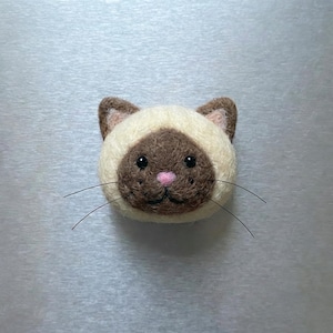 Pu&ograve; includere: Un volto di gatto siamese fatto a mano in feltro. Il gatto ha un muso color crema con segni marroni, un naso rosa e baffi neri. Gli occhi del gatto sono neri e ha un'espressione dolce. Questo gatto in feltro &egrave; un oggetto decorativo.