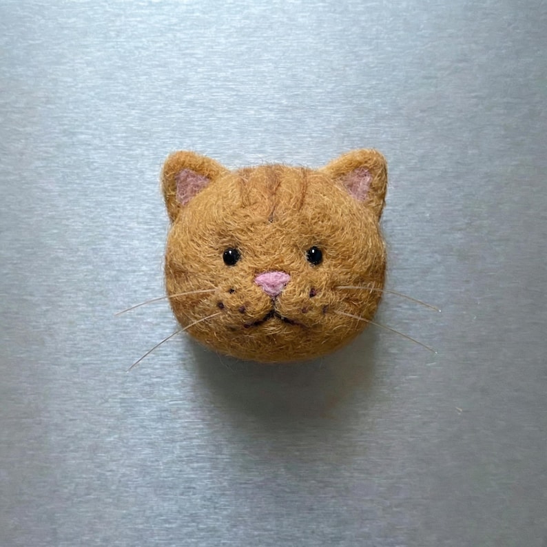 Pu&ograve; includere: Testa di gatto arancione fatta a mano in feltro. Il gatto ha occhi neri, naso rosa e baffi. Le orecchie sono foderate di feltro rosa. Questo oggetto a tema gatto &egrave; probabilmente una spilla o un magnete decorativo.