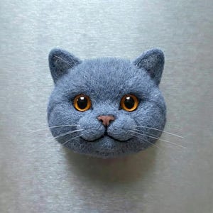 May include: A felted cat head in a shade of blue-gray, featuring large, round orange eyes and a small pink nose. The cat has long white whiskers and a slightly smiling expression, giving it a friendly appearance.