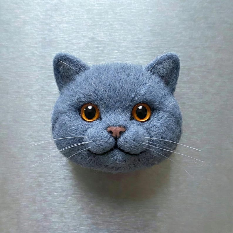 May include: A felted cat head in a shade of blue-gray, featuring large, round orange eyes and a small pink nose. The cat has long white whiskers and a slightly smiling expression, giving it a friendly appearance.