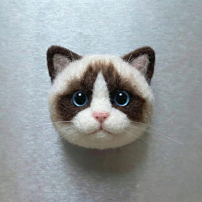 May include: A handcrafted cat face made of felt, featuring a white and brown color scheme. The cat has large blue eyes, a pink nose, and long white whiskers. The cat's face is a unique, handmade item.