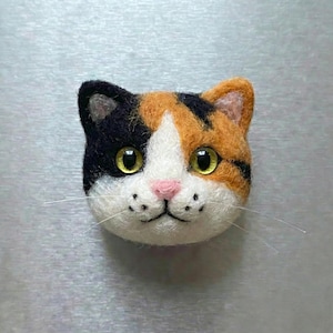 May include: A handcrafted felt cat face with a calico pattern. The cat has black, orange, and white fur, with yellow eyes, a pink nose, and whiskers. The cat's face is a detailed, three-dimensional design.