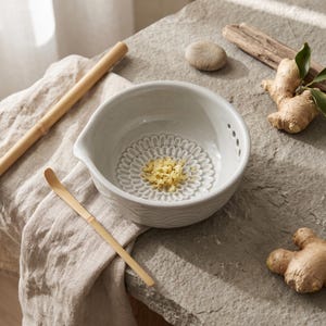 May include: A light gray ceramic ginger grater bowl with a textured, flower-shaped interior base for grating. The bowl features a small pouring spout and a series of drainage holes on one side, containing a small pile of freshly grated ginger.