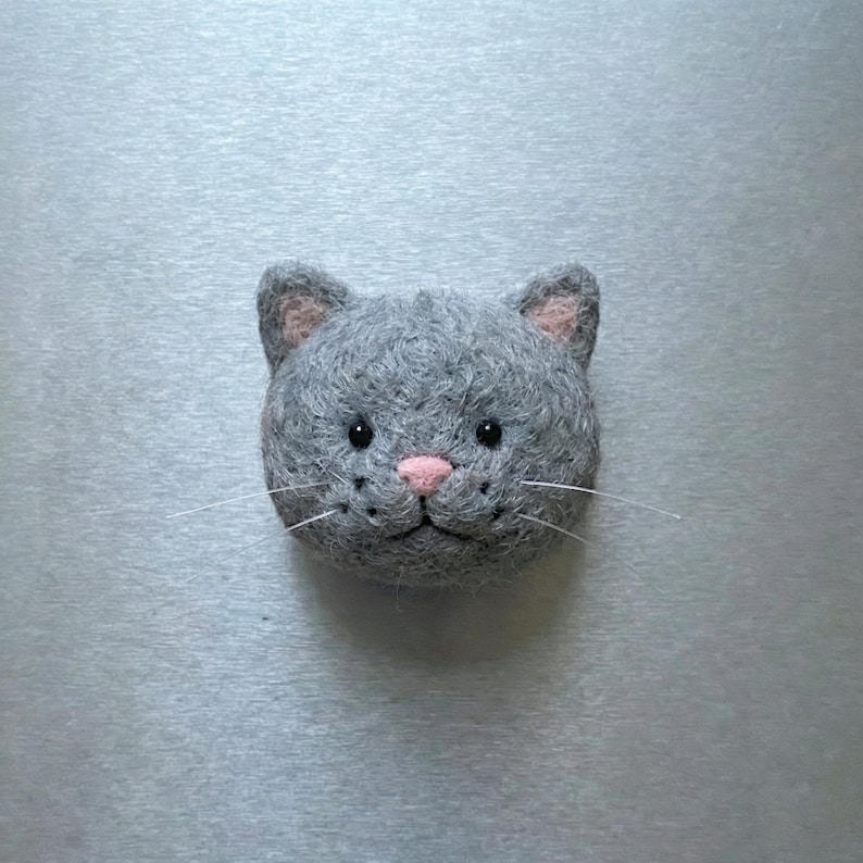 Pu&ograve; includere: Una testa di gatto in feltro grigio con interno orecchie e naso rosa e baffi bianchi. Gli occhi del gatto sono neri e il feltro ha un aspetto strutturato. Il gatto &egrave; centrato nel fotogramma.