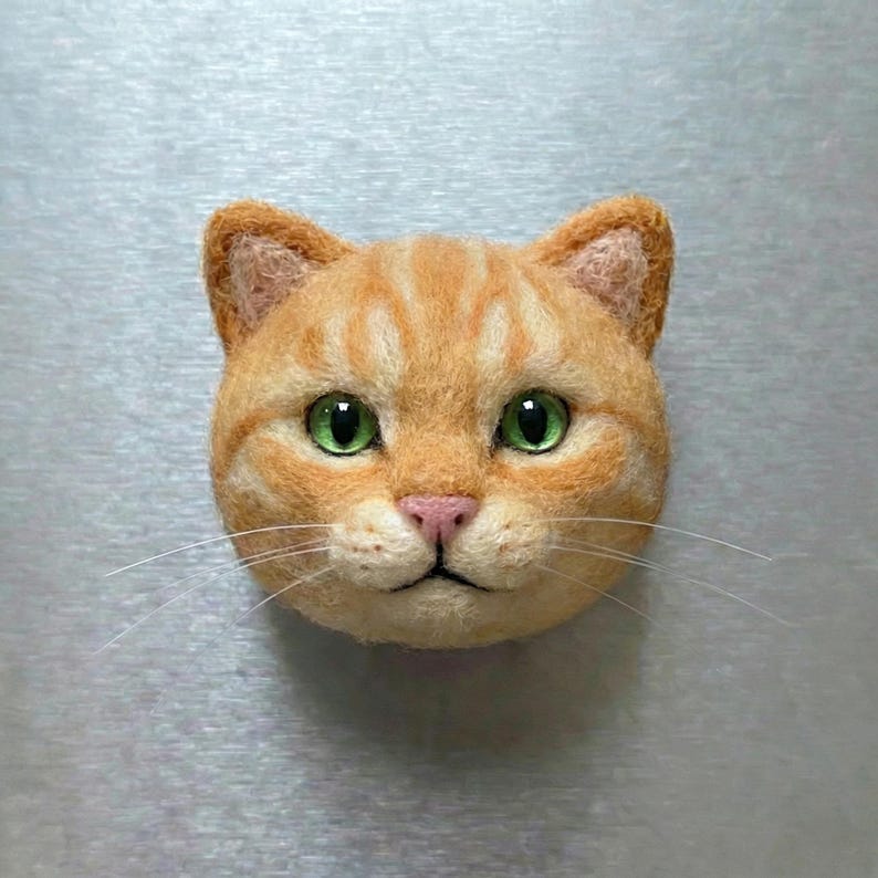 May include: A handcrafted orange tabby cat face with green eyes and a pink nose. The cat's fur is detailed with stripes and texture, and it has long white whiskers. The cat is a decorative item.