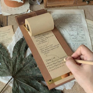 May include: A brown clipboard with a roll of paper and a pencil. The paper roll is attached to the top of the clipboard. The clipboard has a gold-colored metal bar at the bottom. The paper has text that says "Done List" and other project management tasks.