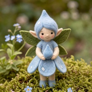 May include: A needle-felted figurine of a small creature with a pointed blue hat, light blue dress, and green leaf-shaped wings. The figure has rosy cheeks, pointed ears, and holds a small blue teardrop shape in its hands, featuring white dots.