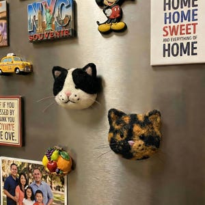 Puede incluir: Dos imanes con forma de gato con texturas afieltradas se exhiben en un refrigerador. Uno es blanco y negro, el otro es carey. Otros imanes incluyen un recuerdo de Nueva York, una figura de Mickey Mouse y un letrero que dice "Home Sweet Home".