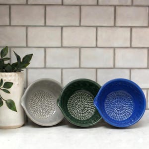 Può includere: Tre ciotole in ceramica grigio chiaro, verde scuro e blu vibrante, ognuna con un motivo strutturato ispirato ai fiori sulla base e un piccolo beccuccio sul lato, disposte in fila accanto a un vaso in ceramica bianca strutturata.