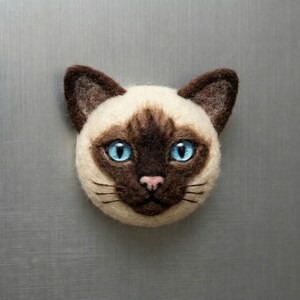 May include: A Siamese cat face made of felt, featuring a cream-colored face with brown markings and large, bright blue eyes. The cat has a pink nose and black whiskers. This handcrafted item is a decorative piece.