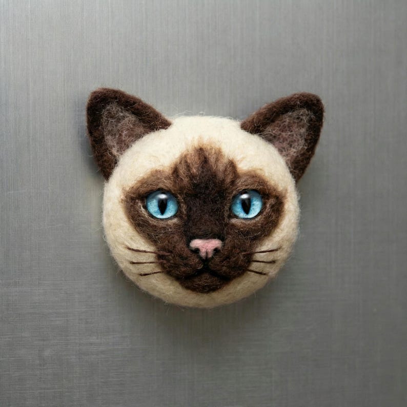 May include: A Siamese cat face made of felt, featuring a cream-colored face with brown markings and large, bright blue eyes. The cat has a pink nose and black whiskers. This handcrafted item is a decorative piece.