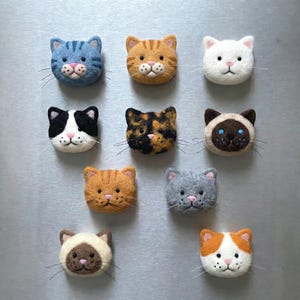 Pode incluir: Uma coleção de ímanes de rosto de gato de feltro feitos à mão em várias cores e padrões. Os ímanes incluem designs de gato azul tigrado, laranja tigrado, branco, preto e branco, tartaruga e siamês. Cada íman tem um nariz rosa e bigodes.