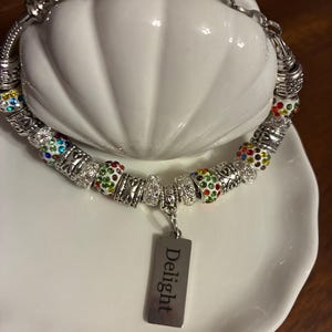 May include: Silver charm bracelet with colorful rhinestone beads and a rectangular charm that reads "Delight". The bracelet is displayed on a white, decorative dish.