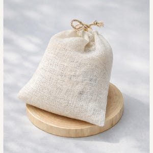 May include: A small, beige burlap bag with a drawstring closure, resting on a round, light-colored wooden coaster. The bag is filled and slightly rounded, suggesting it contains contents. The background is a soft, neutral color.