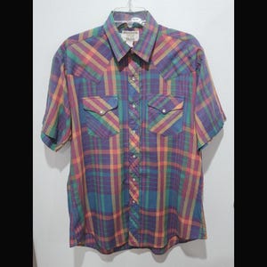 Puede incluir: Una camisa de manga corta con botones y un estampado de cuadros en tonos morado, azul, verde, naranja y amarillo. La camisa tiene dos bolsillos en el pecho con solapas puntiagudas y un cuello. La etiqueta dice "Riddock Shirts".