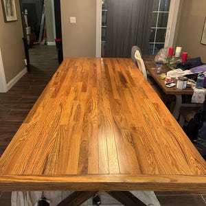 May include: A large, rectangular wooden dining table with a warm, natural wood grain finish. The table has a sturdy, dark brown base and is set in a well-lit dining room. The table is approximately 96 inches long.