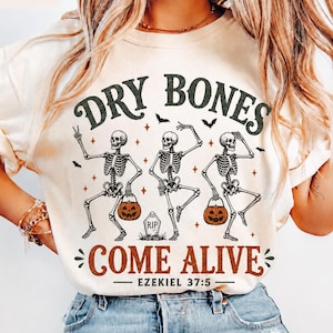 May include: An ivory t-shirt featuring a graphic of three dancing skeletons with pumpkins and bats. The text reads DRY BONES COME ALIVE EZEKIEL 37:5. The design includes a tombstone marked RIP and small star accents in black and orange ink colors.