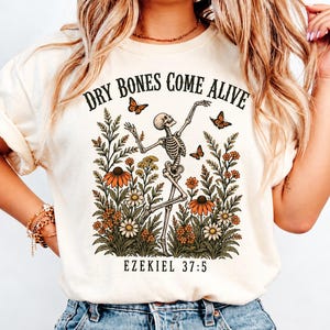 Puede incluir: Una camiseta de color crema con un gráfico de un esqueleto bailando rodeado de flores silvestres naranjas y blancas y mariposas. El texto "DRY BONES COME ALIVE" aparece arriba y "EZEKIEL 37:5" abajo en una fuente serif negra.