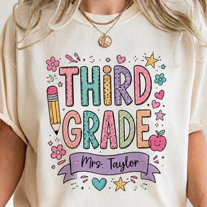 May include: A cream-colored t-shirt featuring the text THIRD GRADE in colorful, patterned block letters. The design includes a pencil, an apple, flowers, and stars. A purple banner below the text displays the name Mrs. Taylor in script.