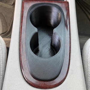 May include: A black dual cup holder insert with a textured finish, designed to fit into a vehicle center console. The insert features two circular openings and is surrounded by a dark reddish-brown wood grain trim piece within a light gray console.