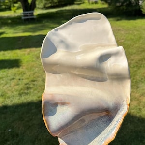 May include: A handcrafted, organic-shaped ceramic serving dish. The dish features a creamy white interior with subtle brown and blue accents along the edges. The exterior has a textured, undulating surface with a golden-brown rim. The dish is held in a person's hand.