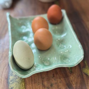 May include: A light green ceramic egg holder with four eggs inside. Two of the eggs are brown and two are white.