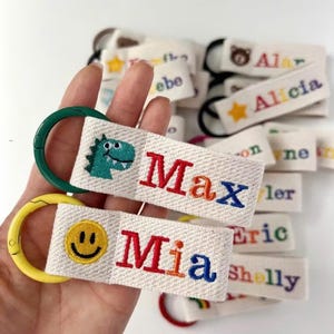 May include: Embroidered fabric keyrings with names and designs. One keyring has a green dinosaur and the name "Max" in rainbow colours. Another has a yellow smiley face and the name "Mia". Other keyrings are visible.